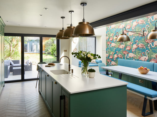 A modern kitchen with a green kitchen island, bronze pendant lights and flamingo patterned wallpaper